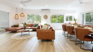 Un vaste espace de vie comprenant un canapé en cuir marron, une table basse ronde et une table à manger avec des chaises. On y trouve également des luminaires suspendus, un ventilateur de plafond et de grandes fenêtres offrant une vue sur la verdure environnante.