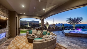 A covered outdoor seating area with a curved sofa, a flat-screen TV, and a view of a mountainous landscape.