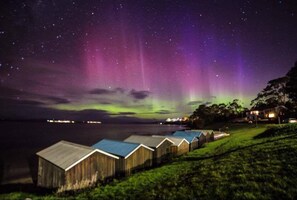Eine Reihe von Holzhütten mit Blick auf das Nordlicht über einem Gewässer.