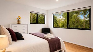 A bedroom with a bed, a nightstand, a chair, and two windows with a view of greenery. - Treetops-Large Home 9km to CBD Sleeps 10. (Taroona)