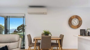 A modern living room with a dining area, a large wooden clock, and a view through a glass door. - Treetops-Large Home 9km to CBD Sleeps 10. (Taroona)