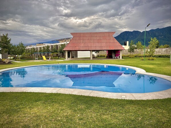 A swimming pool with a red-roofed pavilion in the background, surrounded by green grass and a mountainous landscape.