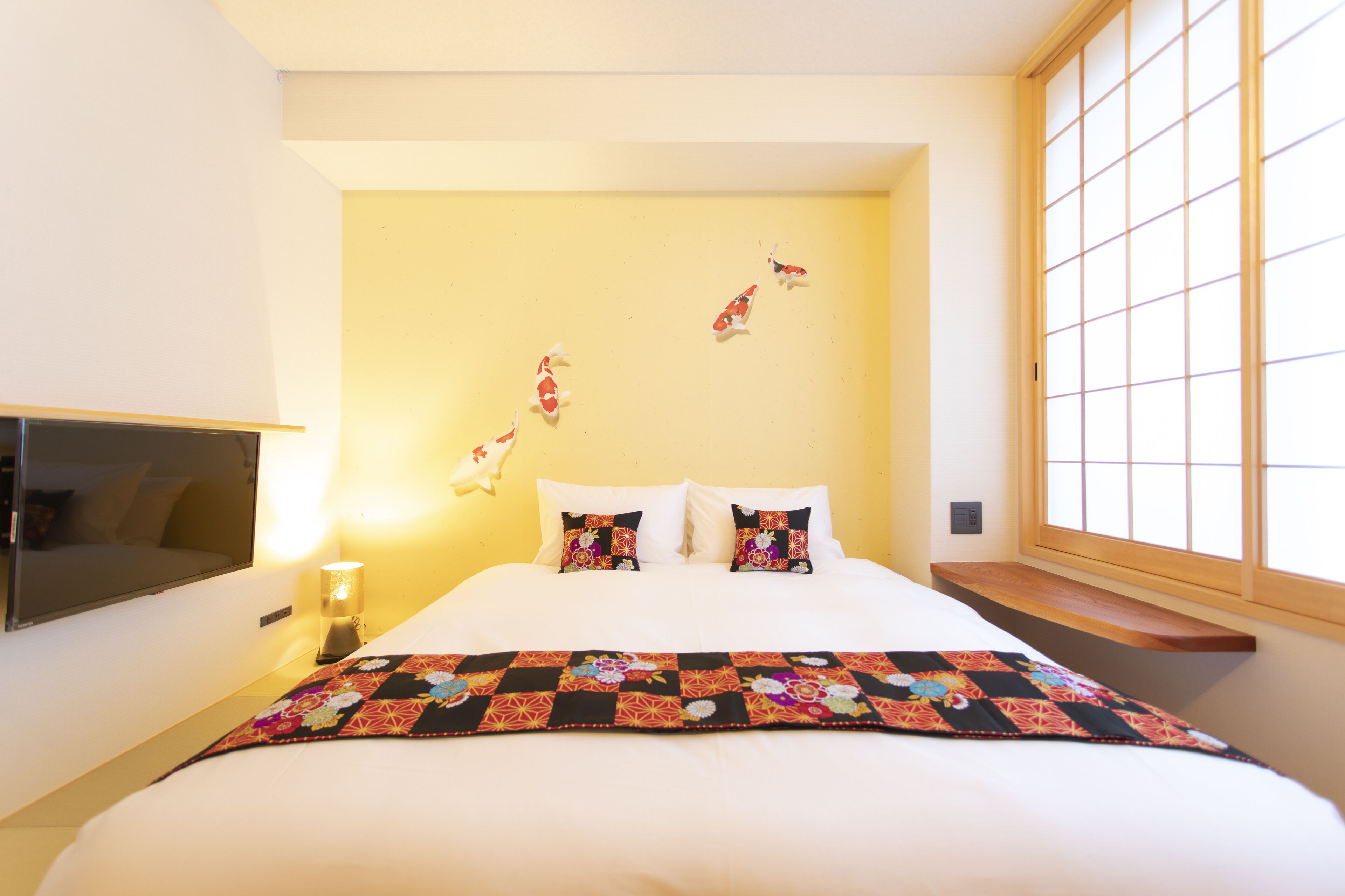 A hotel room with a bed, pillows, a quilt, a bedside table with a lamp, a TV, and a window with a view.