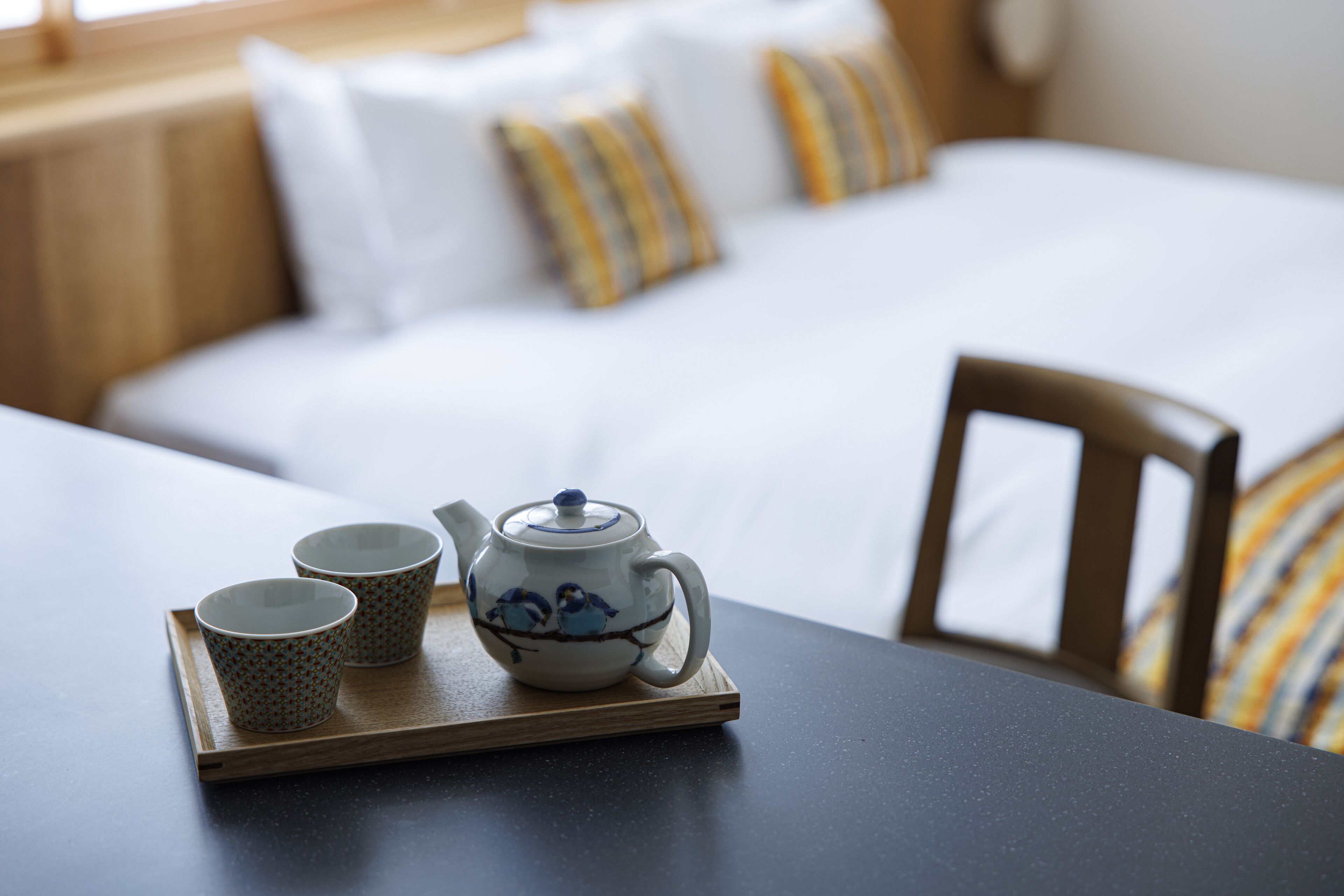 A wooden tray with a teapot and two cups on a table, a bed with white bedding and striped pillows in the background.