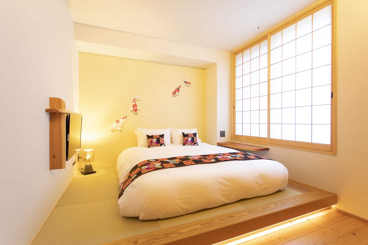 Un dormitorio con una cama, una mesita de noche, una lámpara, almohadas decorativas, una ventana con diseño de cuadrícula y un estante de madera.