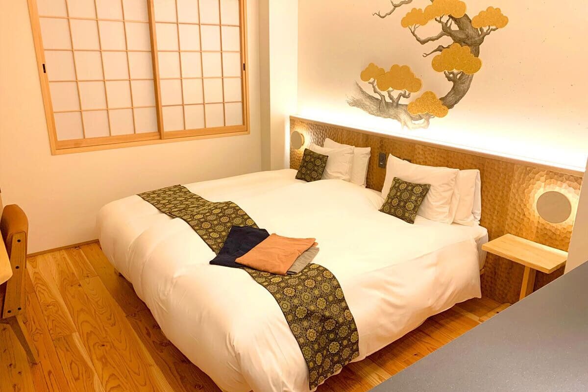 Una cama bien hecha con un plaid estampado, una mesita de noche con espejo y una pared decorada con un cuadro de un árbol.