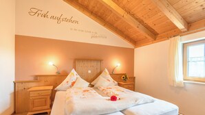 A bedroom with a wooden ceiling, a bed with floral bedding, two bedside tables with lamps, and a window with curtains. - Cornflower apartment 40 sqm (Palling)