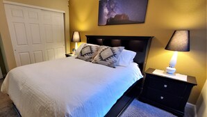 Una cama bien hecha con almohadones estampados, una mesita de noche con lámpara, y un cuadro colgado en la pared que representa un cielo estrellado.