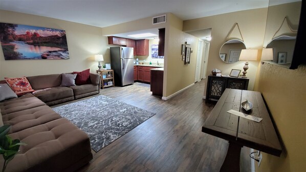 A living room with a brown sofa, a patterned rug, and a painting on the wall. There is a kitchen area with wooden cabinets and a refrigerator.