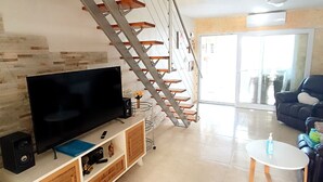 Una sala de estar con televisor, parlantes y una mesa de centro. Una escalera con escalones de madera y barandillas de metal conduce a otro nivel.