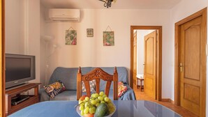 Un salon avec un canapé, un téléviseur, une table à manger ornée de fruits et un couloir menant à une porte.