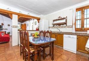 Un comedor con mesa de vidrio, sillas de madera, frutero, cocina con armarios blancos, lavavajillas blanco y ventana con cortinas.