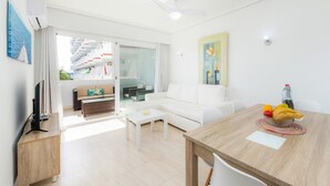 A modern living room with a white sofa, a dining table with fruit, a TV, and a balcony with a view of a building. - Holiday Apartment "Alcudia Sea" with Large Balcony, Pool View & Wi-Fi (Home Office) (Alcudia)