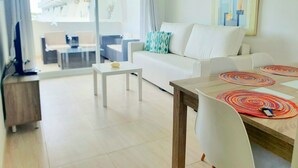 A modern living room with a white sofa, a dining table with colorful placemats, a television, and a large window with a view of buildings outside. - Holiday Apartment "Alcudia Sea" with Large Balcony, Pool View & Wi-Fi (Home Office) (Alcudia)