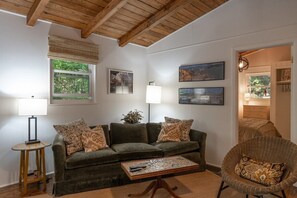A living room with a sofa, a coffee table, a lamp, and a chair. There are framed pictures on the wall and a window with blinds.