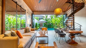 A modern living room with a sofa, coffee table, and dining table, a spiral staircase, and a view of lush greenery outside. - Luxury Private Loft, 2 BR, Canggu With Staff (Canggu)