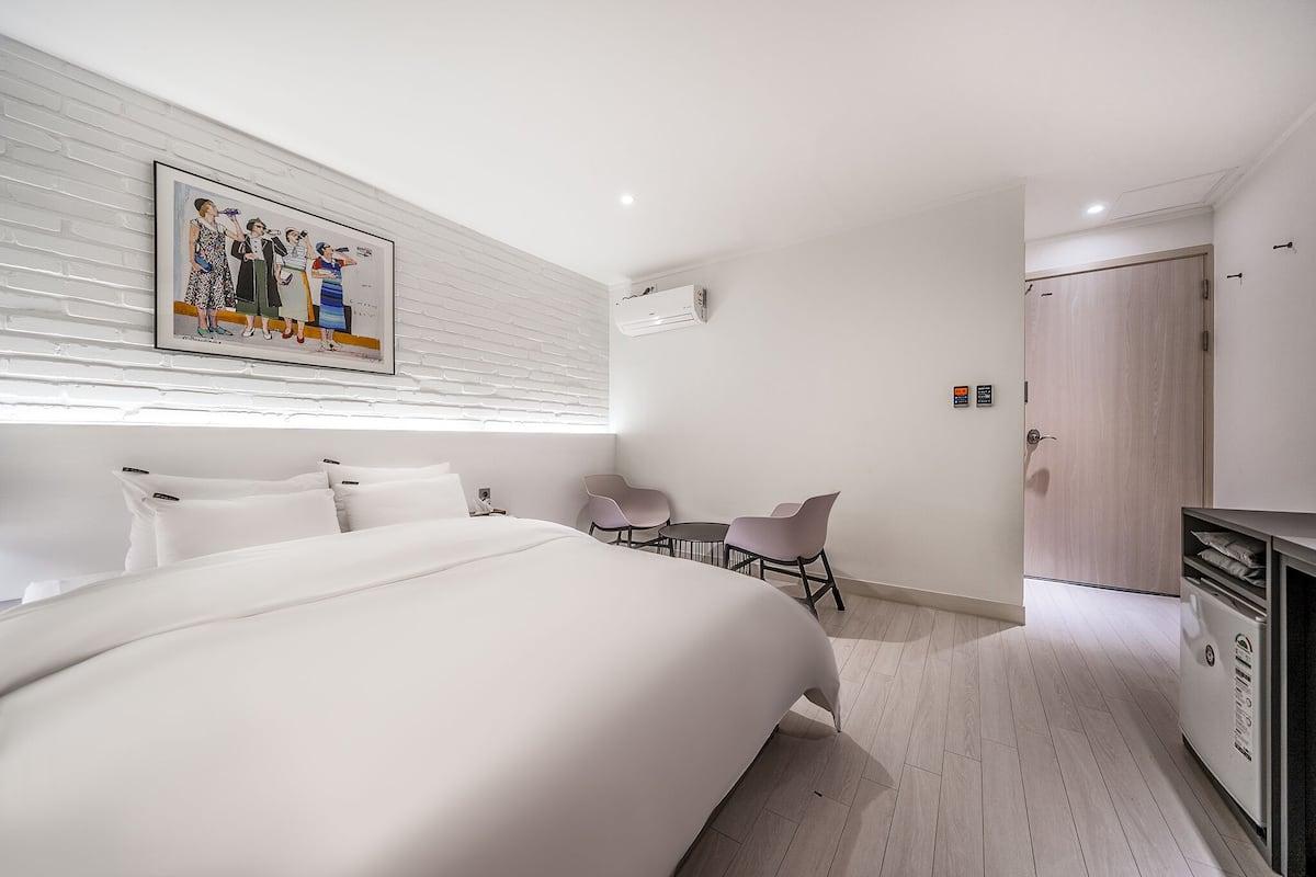 Una habitación de hotel moderna con una cama grande, un escritorio pequeño con dos sillas, una pared de ladrillo blanco y una puerta con acabado en madera clara.