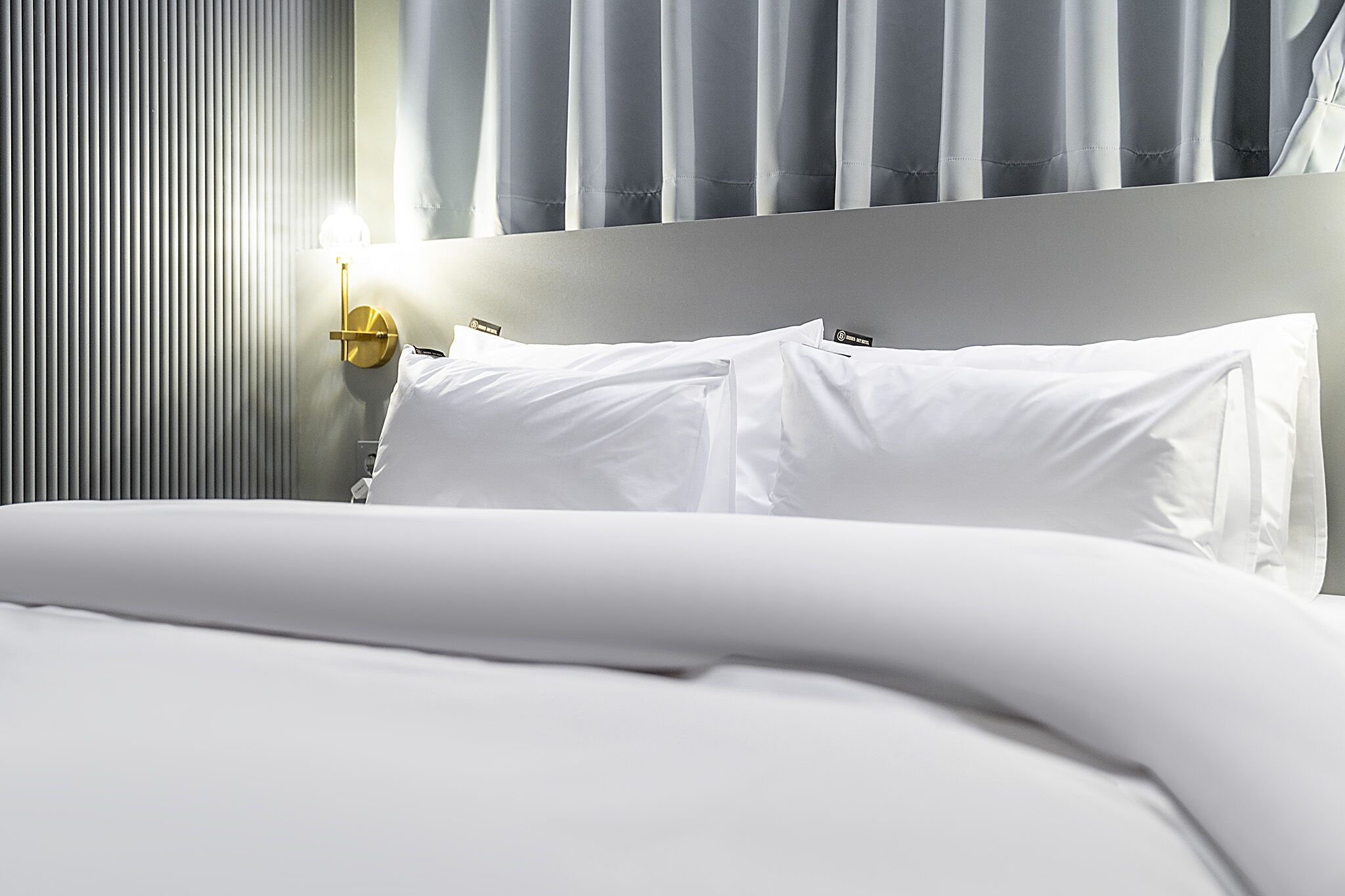 Un lit bien fait, avec du linge de lit blanc et des oreillers, une table de chevet avec une lampe, et une fenêtre avec des rideaux.