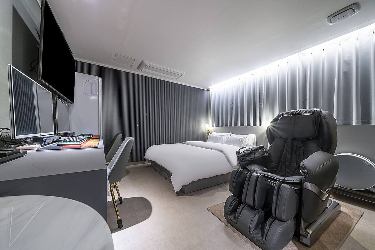 Habitación de hotel moderna con cama, sillón de masajes, escritorio con ordenador y un amplio ventanal con cortinas.