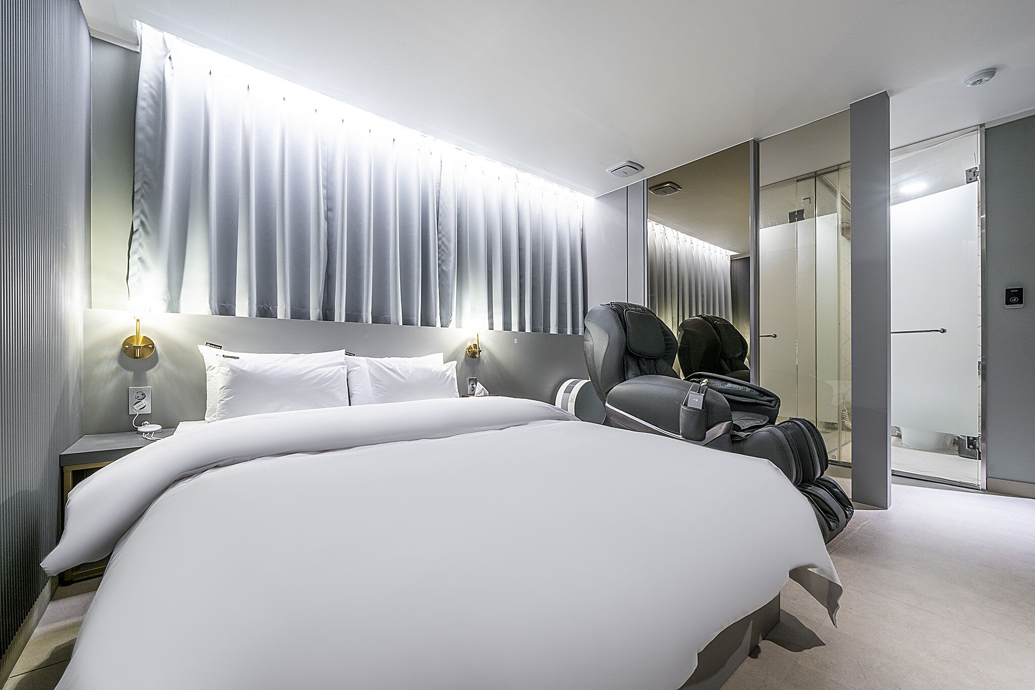 Una habitación de hotel moderna con una cama grande, un área de estar con un sillón de masaje y un espejo grande.