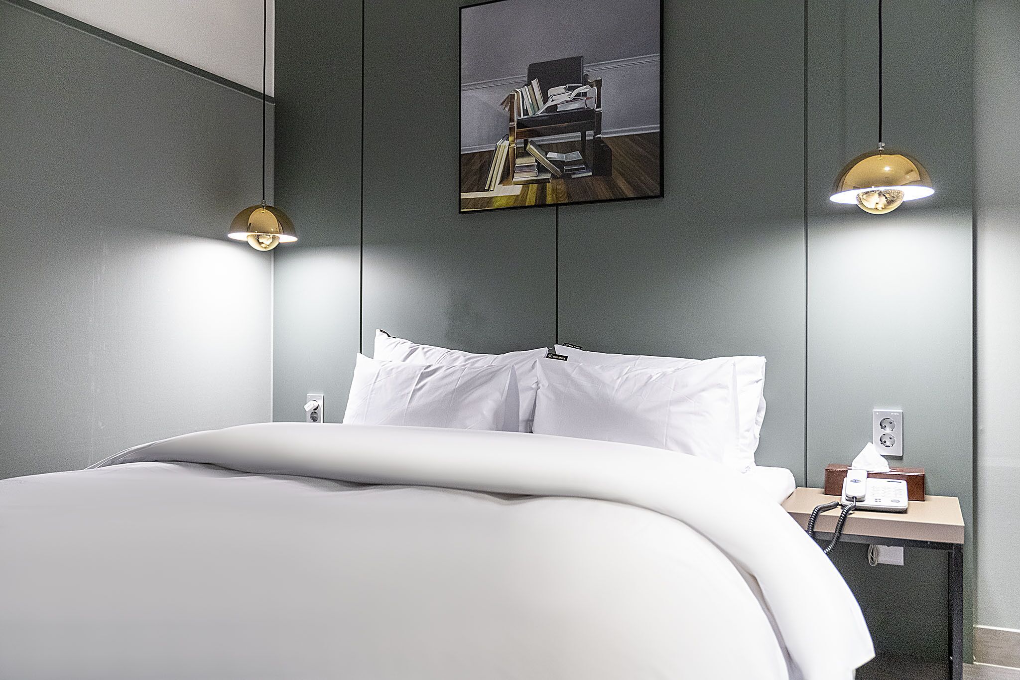 A modern hotel room with a neatly made bed, a bedside table with a lamp, and a painting on the wall.