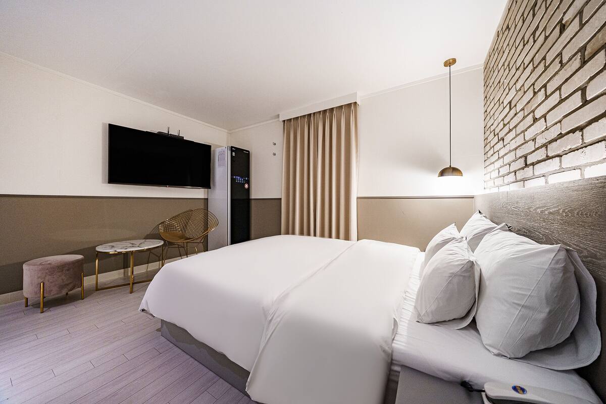 Habitación de hotel con una cama grande, un televisor de pantalla plana en la pared, una mesita redonda pequeña, un banco acolchado y una pared con diseño de ladrillo.