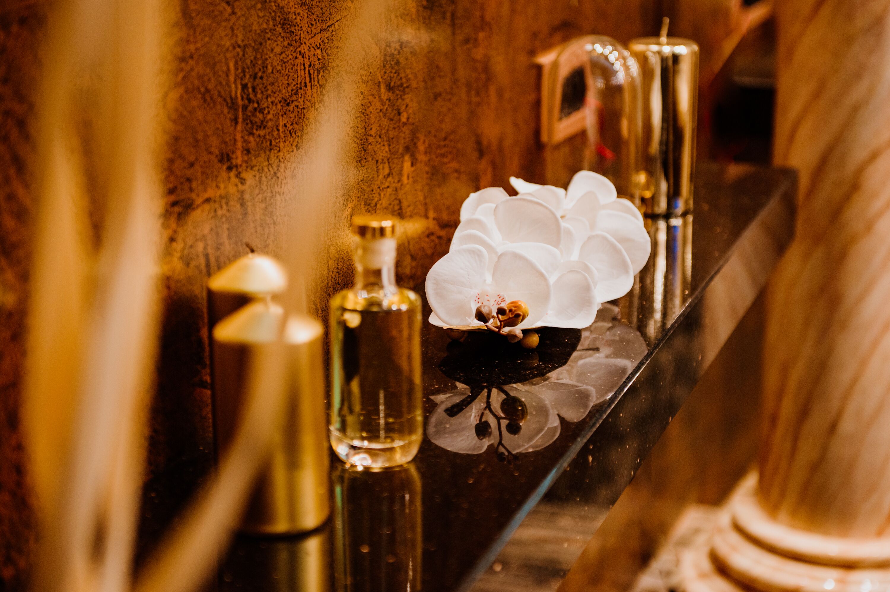 A bathroom vanity with a mirror, a white orchid, and two gold-colored bottles.