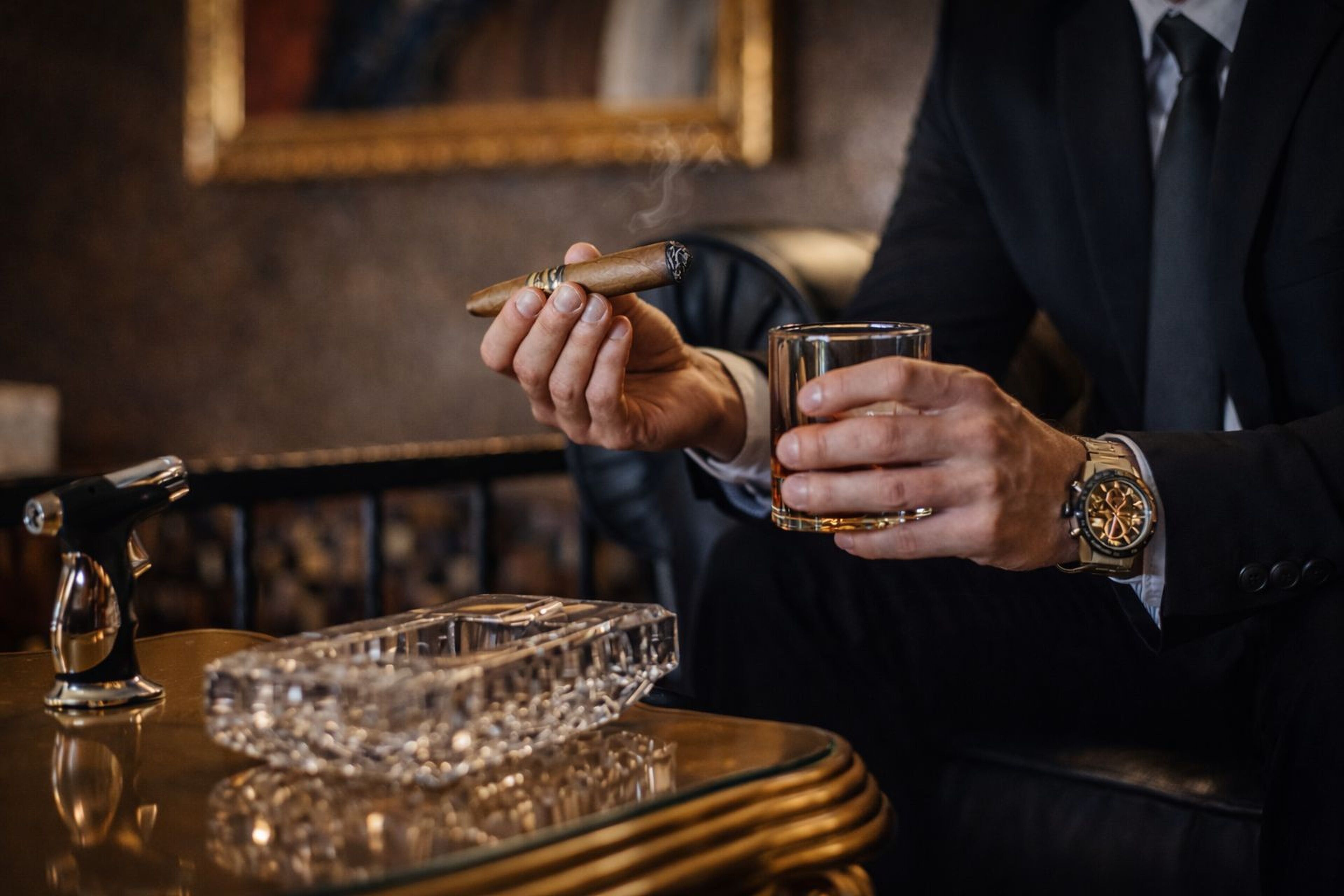 Una persona sostiene un puro y una copa de whisky, con un posavasos de cristal sobre una mesa.