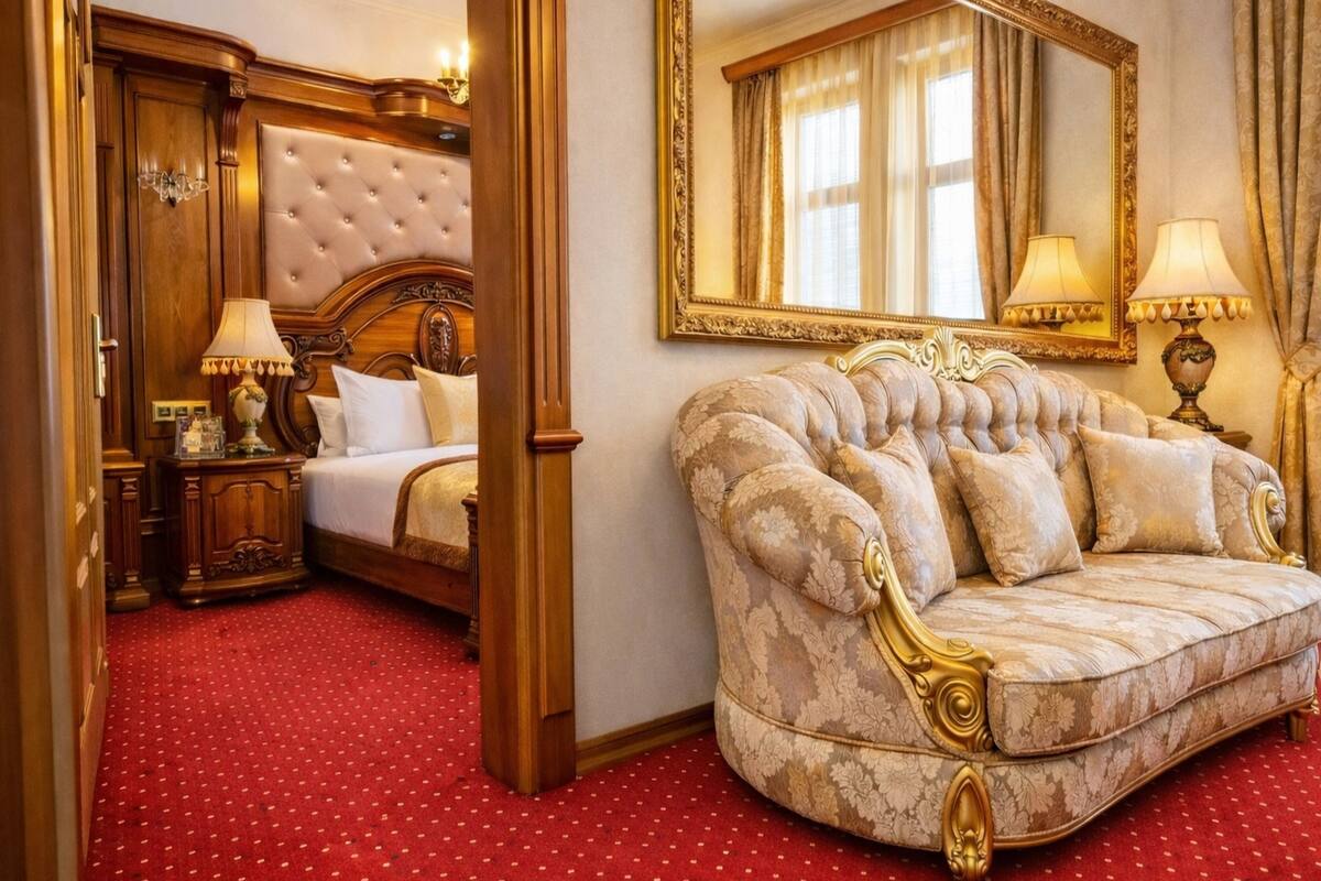 A luxurious hotel room with a tufted headboard bed, a plush sofa, a large ornate mirror, and a red carpeted floor.
