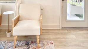 Quarto com cadeira branca, pequena mesa redonda, tapete estampado, porta com vista para uma varanda e chapéu fixado na parede.