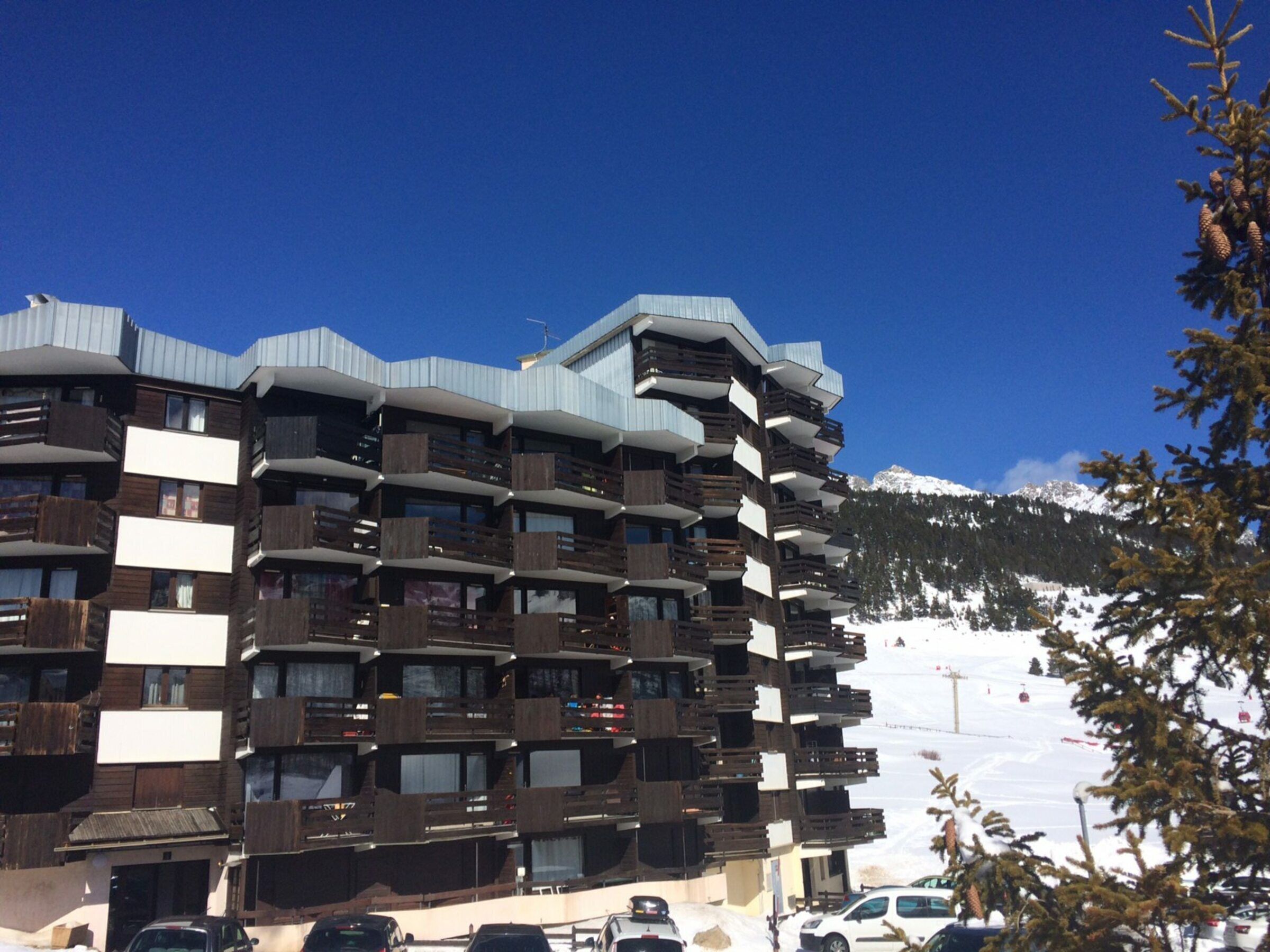 Un hotel de varios pisos con balcones, una pista de esquí al fondo y un cielo azul despejado.