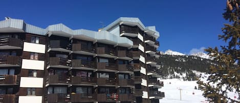 Un hotel de varios pisos con balcones, una pista de esquí al fondo y un cielo azul despejado.