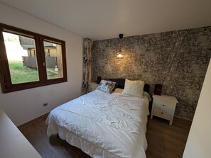 A bedroom with a large bed, a nightstand, a wall-mounted lamp, and a window with a view of the outside.
