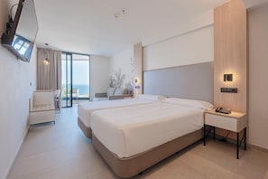 A modern hotel room with two beds, a TV, a desk, and a view of the sea.