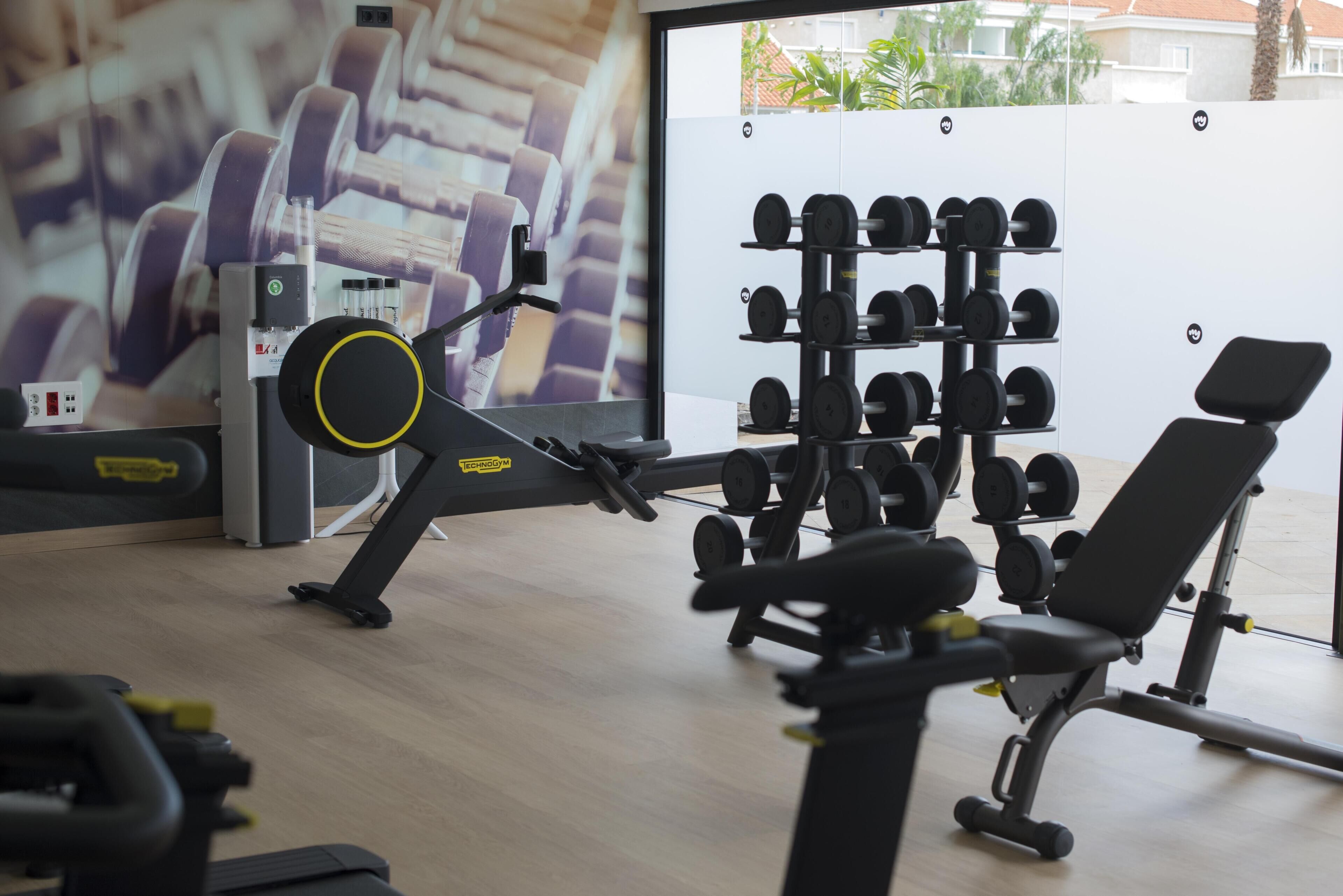 Una palestra con attrezzature per esercizi, una rastrelliera per manubri a parete e una finestra con vista su edifici e alberi.