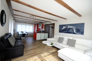 Ein modernes Wohnzimmer mit einem weißen Sofa, einem schwarzen Ledersessel und einem Essbereich, im Hintergrund eine rote Küche.