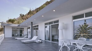 Ein moderner Außenbereich mit Loungemöbeln in Weiß, darunter Liegestühle und ein Tisch, sowie große Fenster mit Blick auf die umliegende Landschaft.
