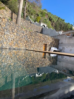 A stone wall with a small window, a clear pool reflecting the sky, and a palm tree. - CARRE NOIR | House45 | Design&Nature (La Orotava)