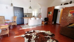 Un salón espacioso con un gran tapete de cuero, un comedor con sillas, una cocina al fondo y una pared de ladrillo decorativa.