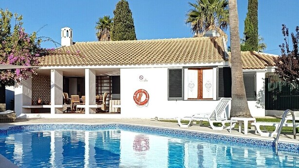 Un área junto a la piscina con una pileta revestida, sillas de descanso y un edificio blanco con techo de tejas.