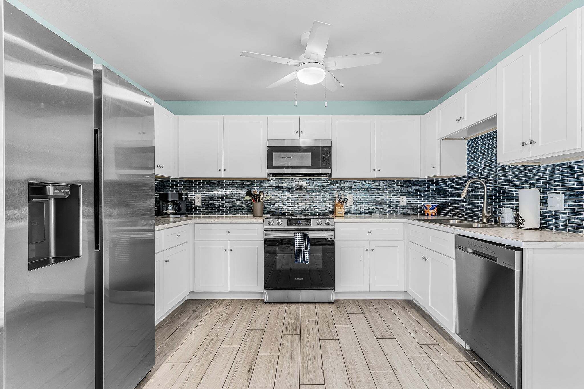 A modern kitchen with stainless steel appliances, white cabinetry, a blue-tiled backsplash, and a ceiling fan.