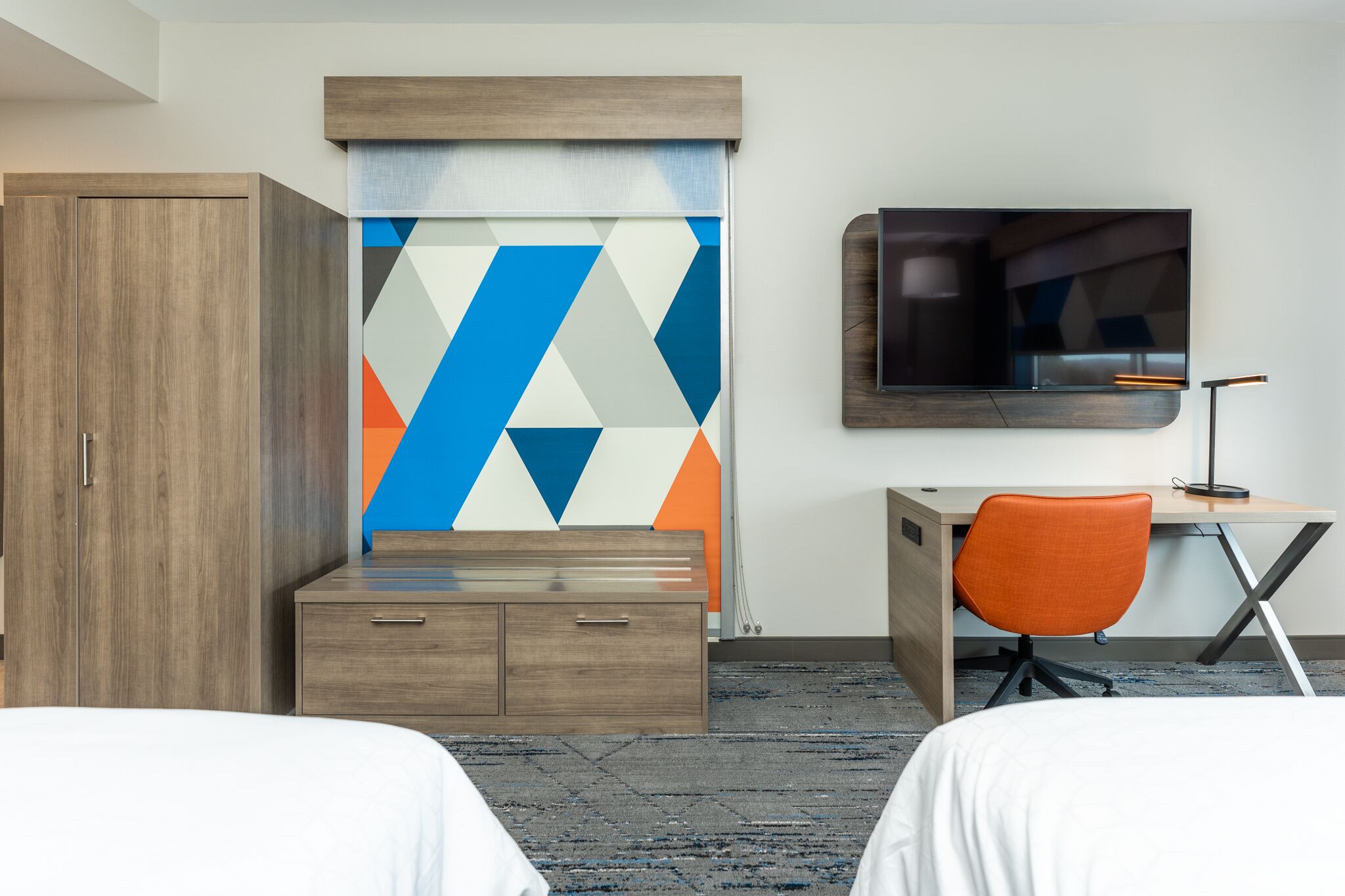 A hotel room with two beds, a desk with an orange chair, a TV, and a wardrobe.