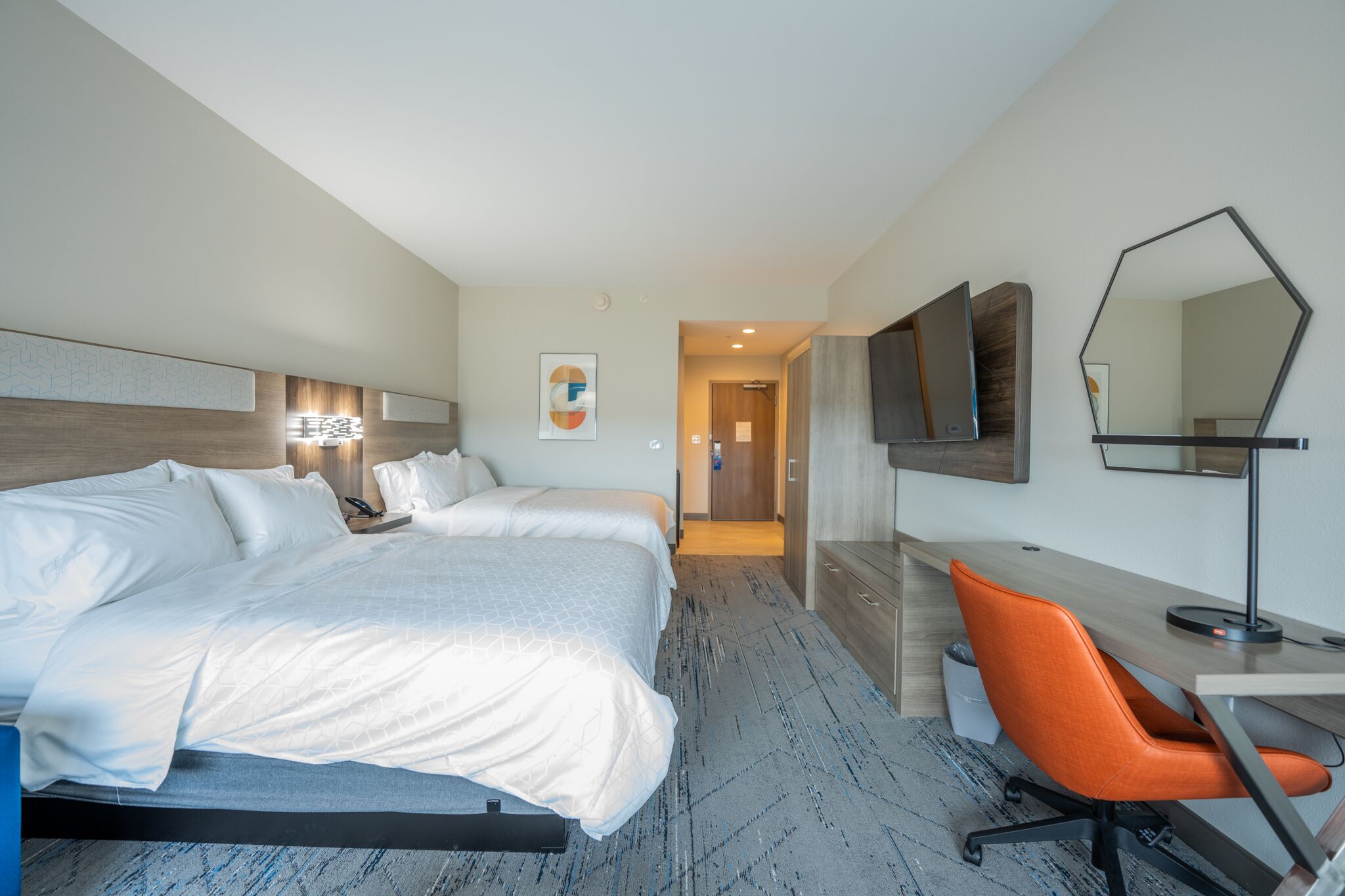 A hotel room with two beds, a desk, an orange chair, a TV, and a mirror.