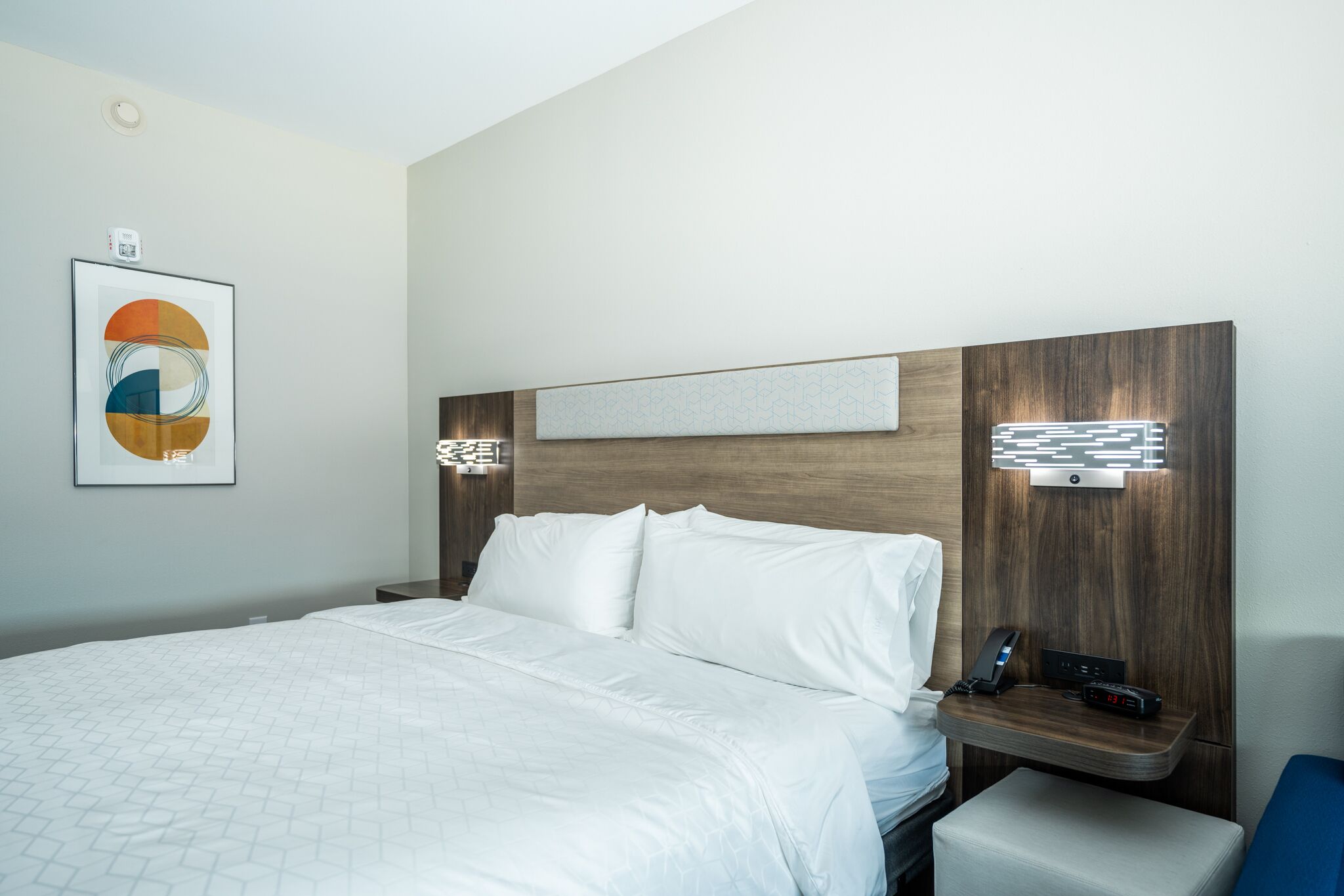 Een hotelkamer met een groot bed, een nachtkastje met een telefoon en oplader, een wandlamp en een kunstwerk in een lijst boven het bed.