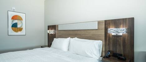 A hotel room with a large bed, a bedside table with a phone and charger, a wall-mounted light, and a framed artwork above the bed.