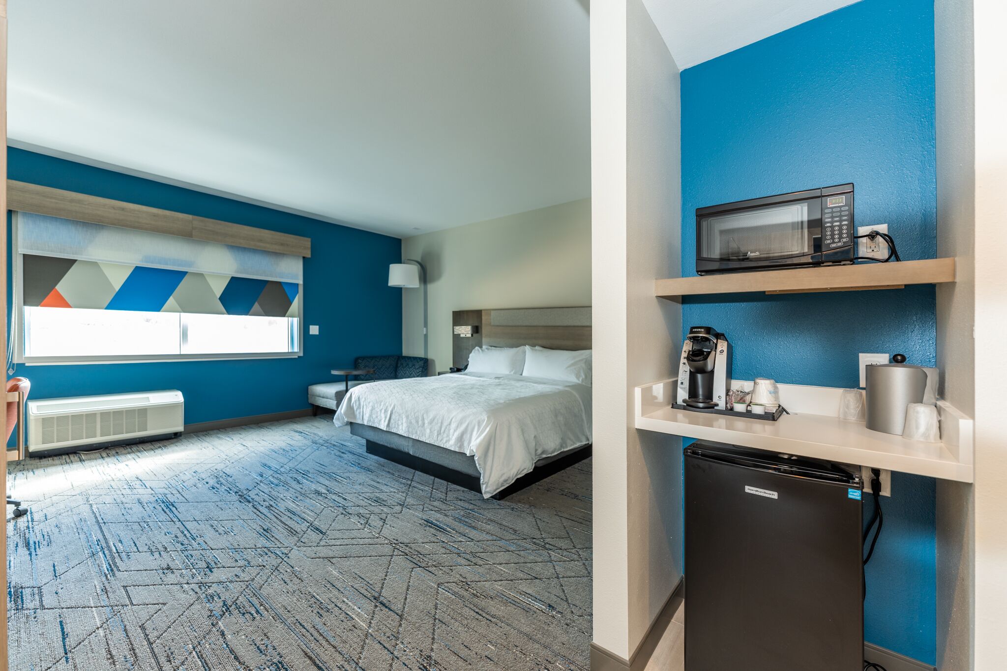 Habitación de hotel con cama, escritorio, microondas, cafetera y refrigerador.