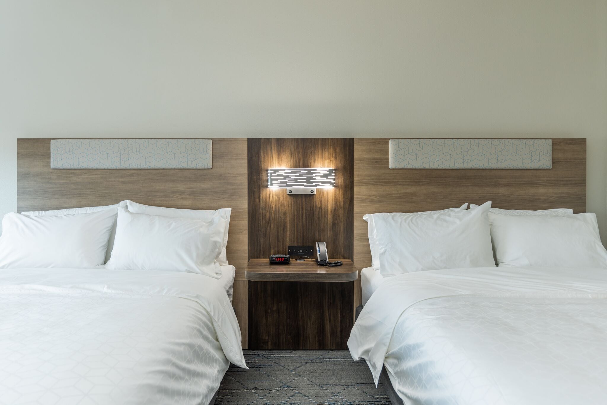 Quarto de hotel com duas camas, uma cabeceira de madeira, mesa de cabeceira com telefone e relógio, e luminária fixada na parede.