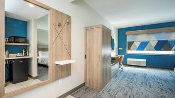 Une chambre d’hôtel moderne dotée d’un grand miroir, d’une armoire en bois, d’une petite kitchenette et d’un lit avec du linge de lit blanc.