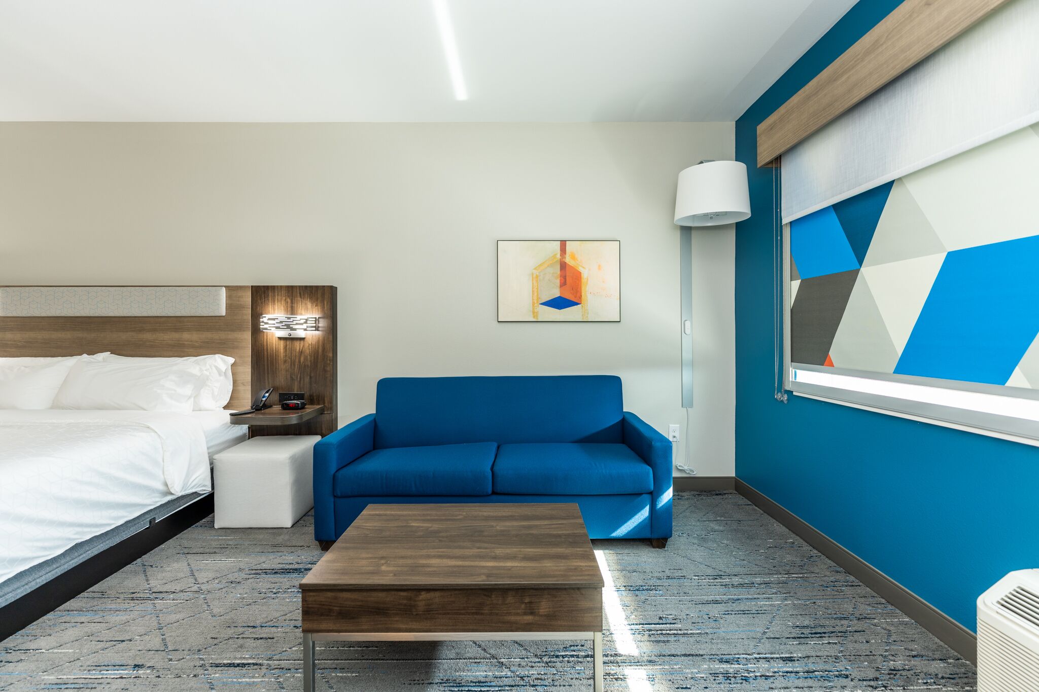 A modern hotel room with a bed, a blue sofa, a wooden coffee table, and a geometric wall design.