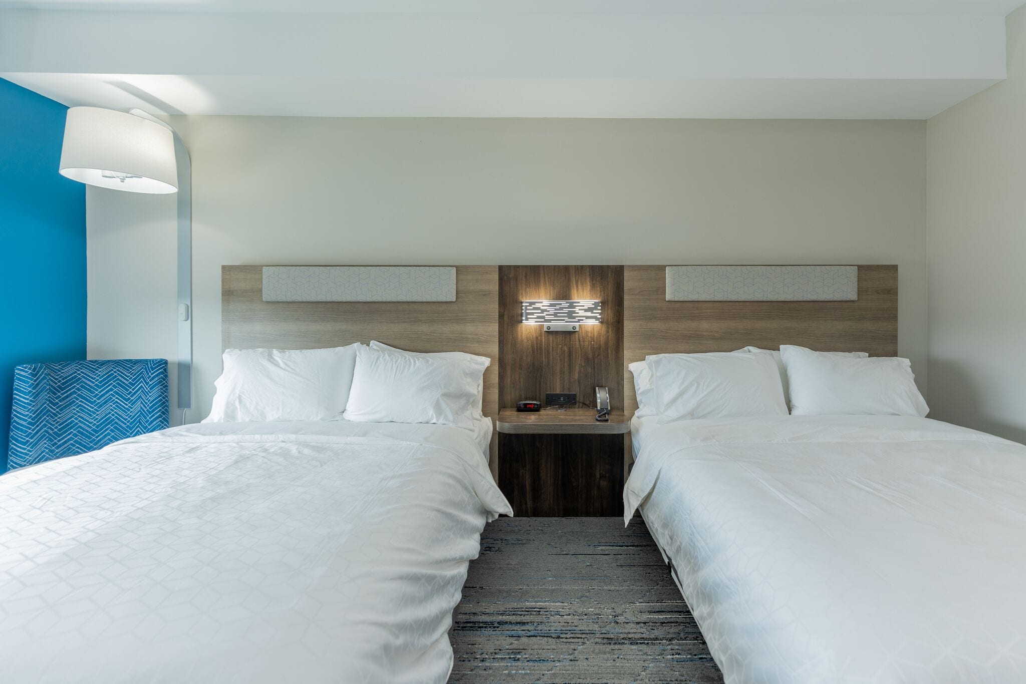 A hotel room with two beds, a wooden headboard, a nightstand with a phone and a clock, and a blue accent wall.